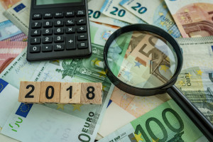financial tax concept as magnifying glass on pile of euro banknotes, calculator and number 2018 on wooden block